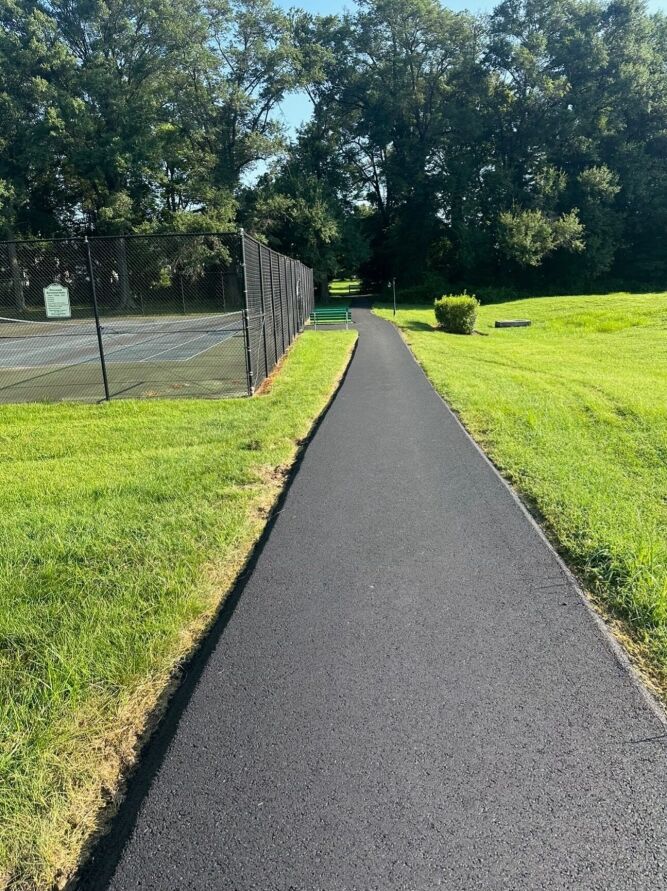 A newly paved asphalt path, expertly crafted by a commercial paving contractor, runs alongside a tennis court and grassy field, bordered by trees in the background on a sunny day.