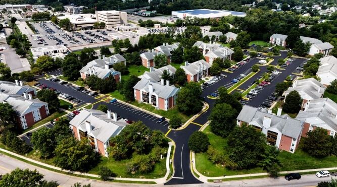 Aerial view of a residential apartment complex with multiple buildings, parking lots crafted by a commercial paving contractor, and surrounding greenery, located near a commercial area.