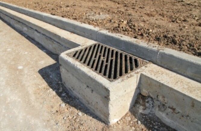 A rectangular storm drain with a metal grate is built into the concrete curb along the edge of a dirt and gravel road, expertly installed by a commercial paving contractor.