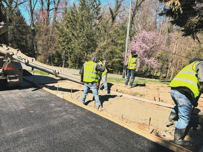 Four construction workers in yellow safety vests pour and spread concrete from a truck onto a prepared section of road, completing concrete sidewalk repair amid trees and a flowering tree in the background.