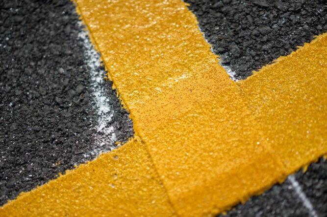 Close-up of a textured yellow roadway thermoplastic marking painted on dark asphalt, forming a right angle with some white line visible in the background.
