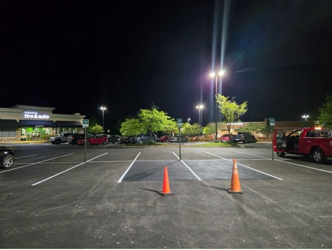 A mostly empty parking lot at night, maintained by a commercial paving contractor, with two orange cones blocking a space and a few cars parked near a row of trees under streetlights.
