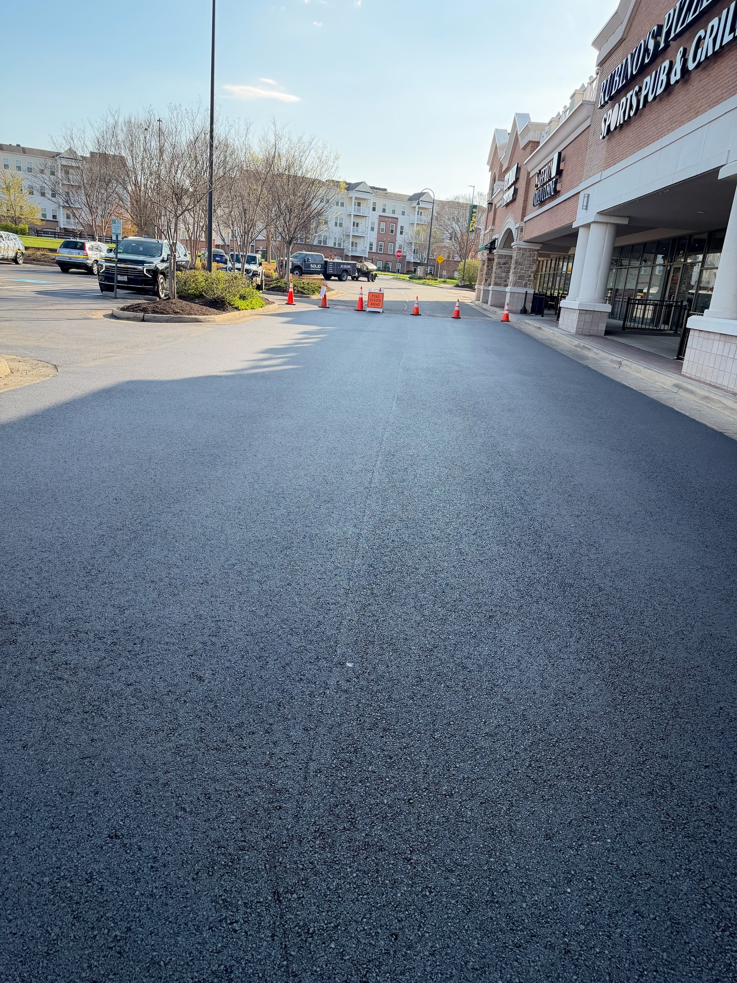 Freshly paved asphalt road in front of a strip mall, with traffic cones blocking the entrance, cars parked along the sides, and nearby concrete sidewalk repair in progress.