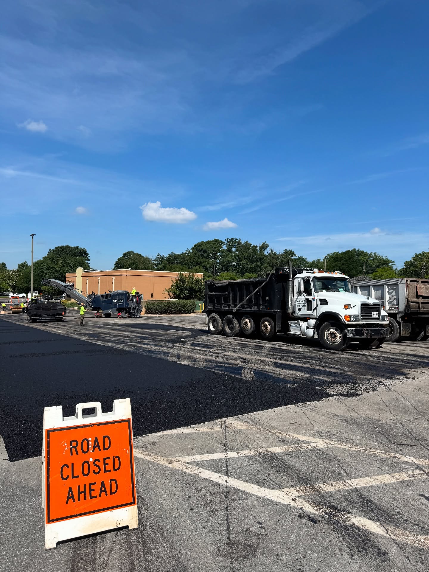 A construction crew is resurfacing a road with trucks and asphalt equipment. A Road Closed Ahead sign is placed in the foreground.