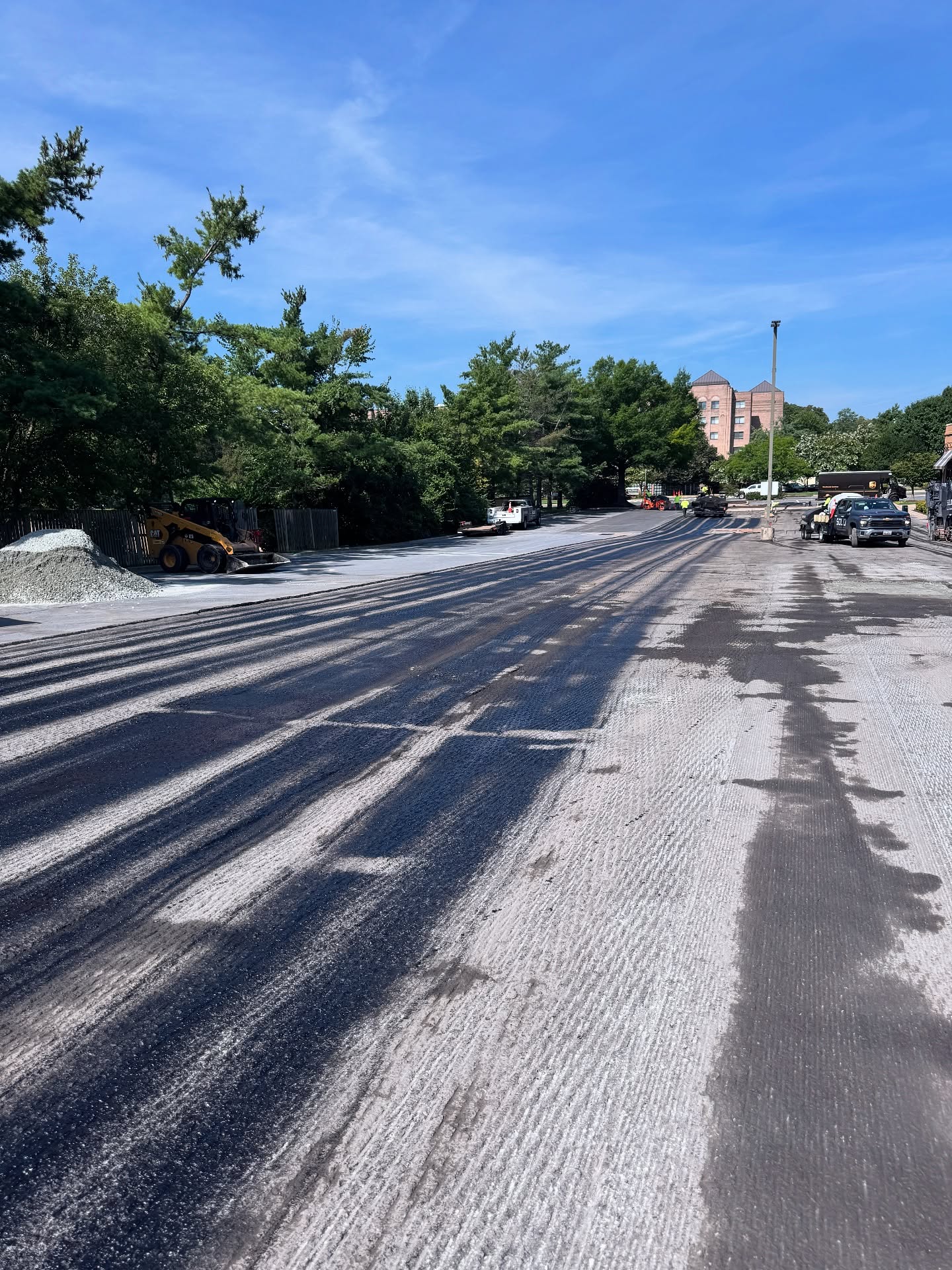A partially resurfaced road with construction vehicles and equipment visible; trees line the sides and buildings are seen in the background under a clear sky.