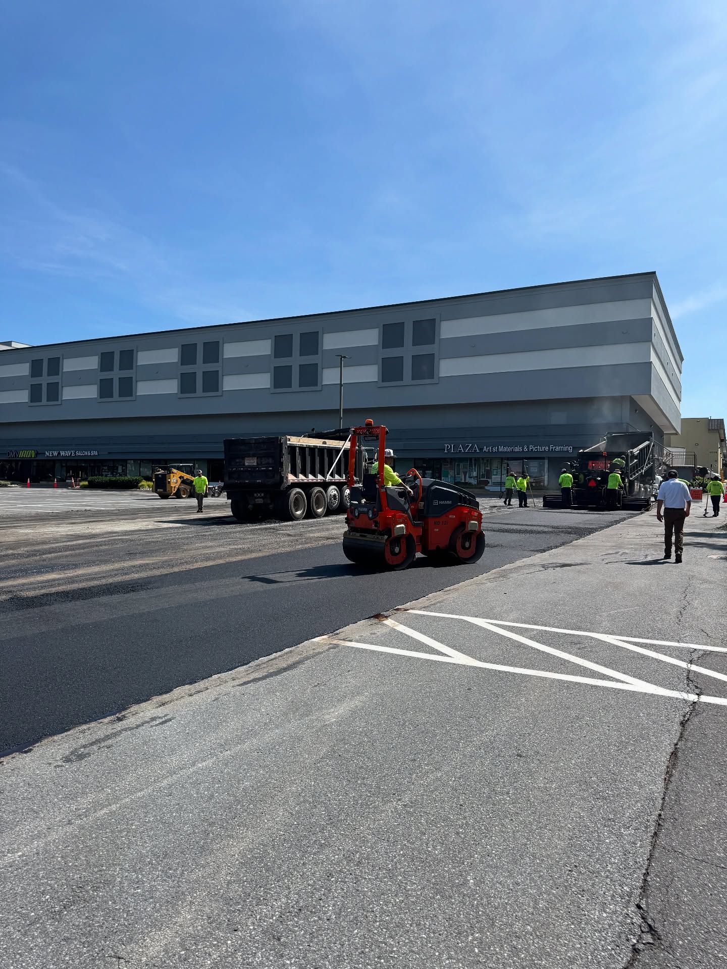 Workers and construction vehicles are resurfacing a parking lot in front of a large commercial building under a clear blue sky.