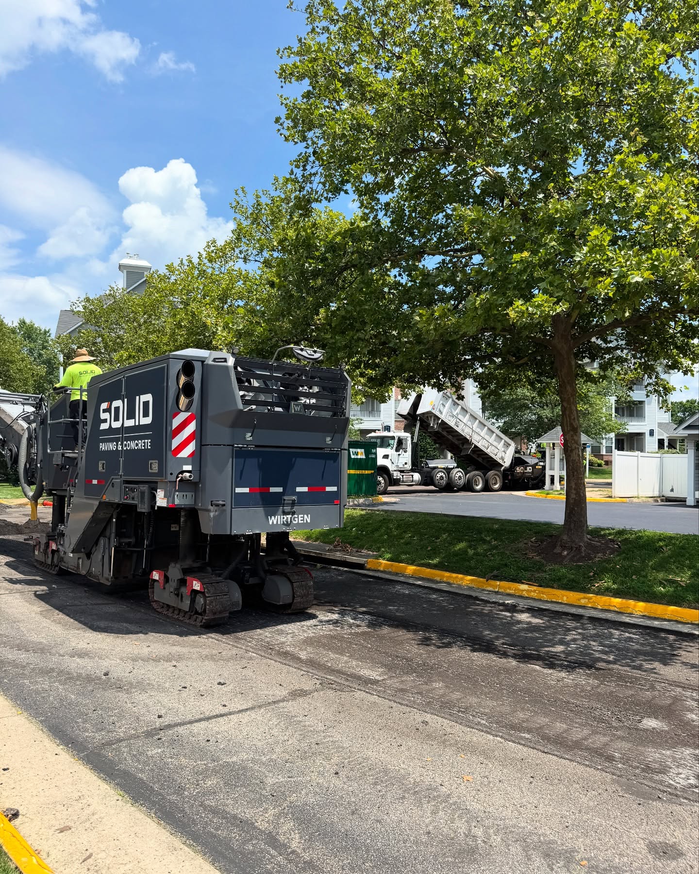 A road milling machine labeled Solid Paving & Concrete works on a street as a dump truck unloads materials nearby; trees and houses are in the background.