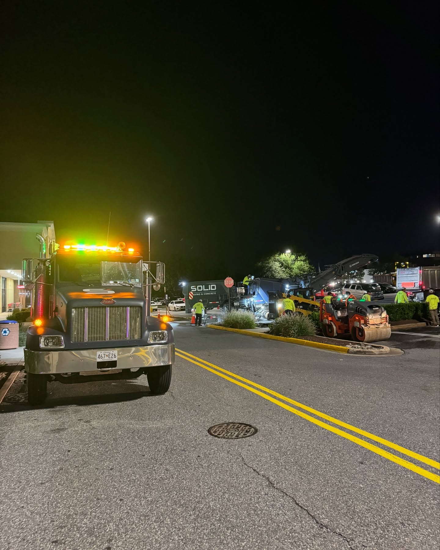 A nighttime roadwork scene with construction vehicles, workers in safety vests, and equipment under streetlights. A large truck with flashing lights is parked on the left side of the road.