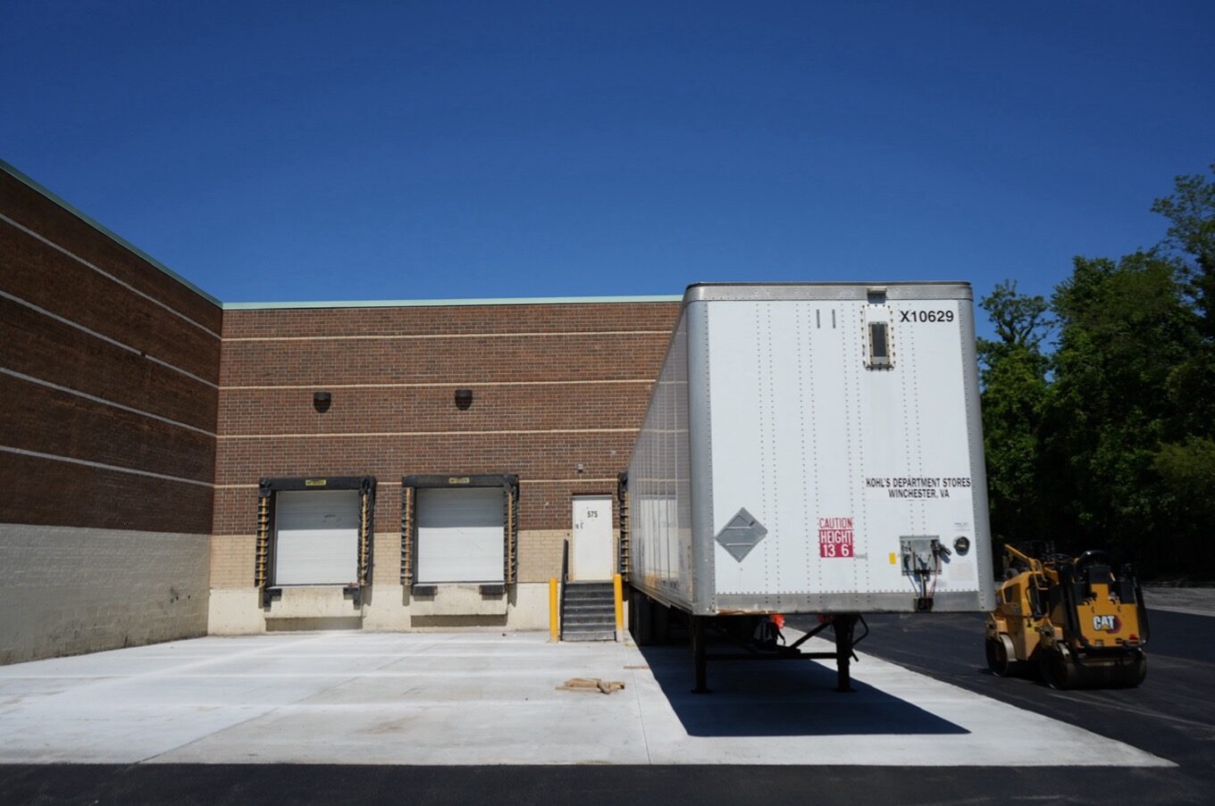 A semi-trailer is parked at a loading dock outside a brick industrial building with two empty loading bays, freshly maintained by a commercial paving contractor, and a forklift nearby.