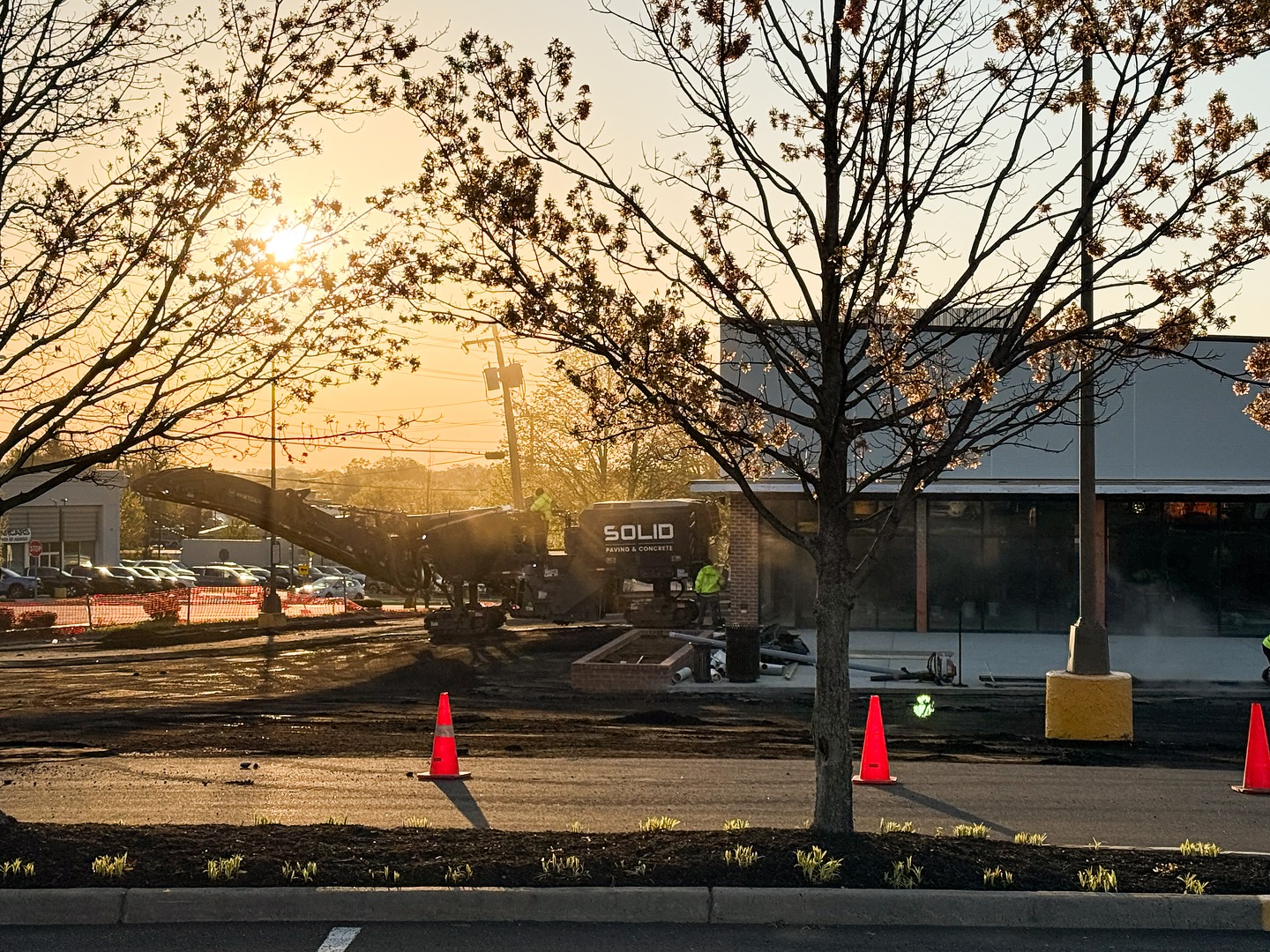 Workers operate heavy machinery at a construction site outside a commercial building at sunset, focusing on concrete sidewalk repair, with orange cones marking the area and trees in the foreground.