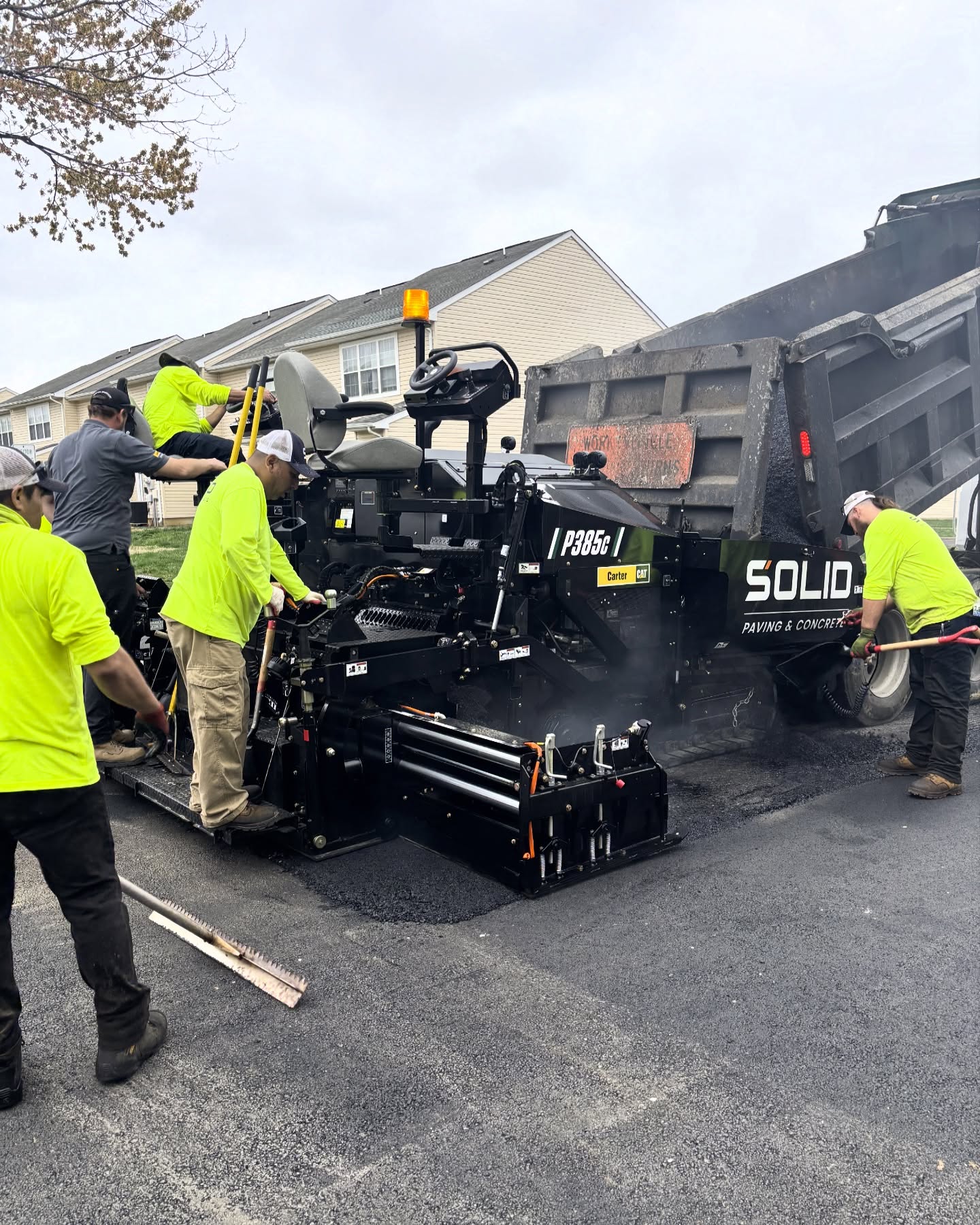 A crew of workers in neon shirts operates a paving machine and dump truck as they lay new asphalt on a residential street.
