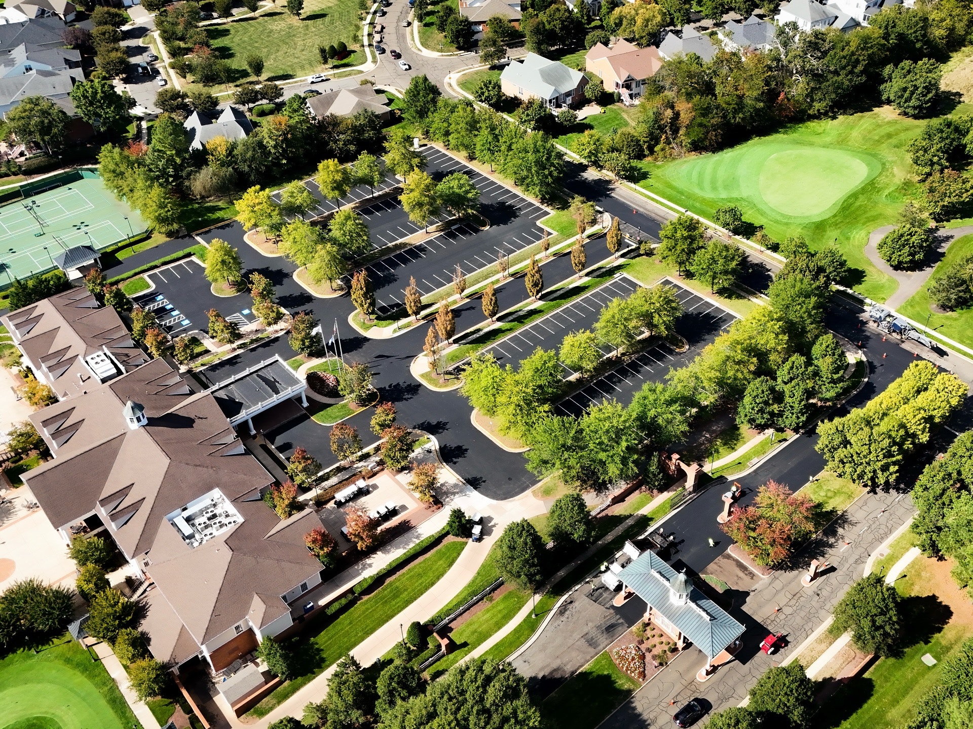 Aerial view of a suburban complex with parking lots, tennis courts, landscaped greenery, and nearby residential houses.